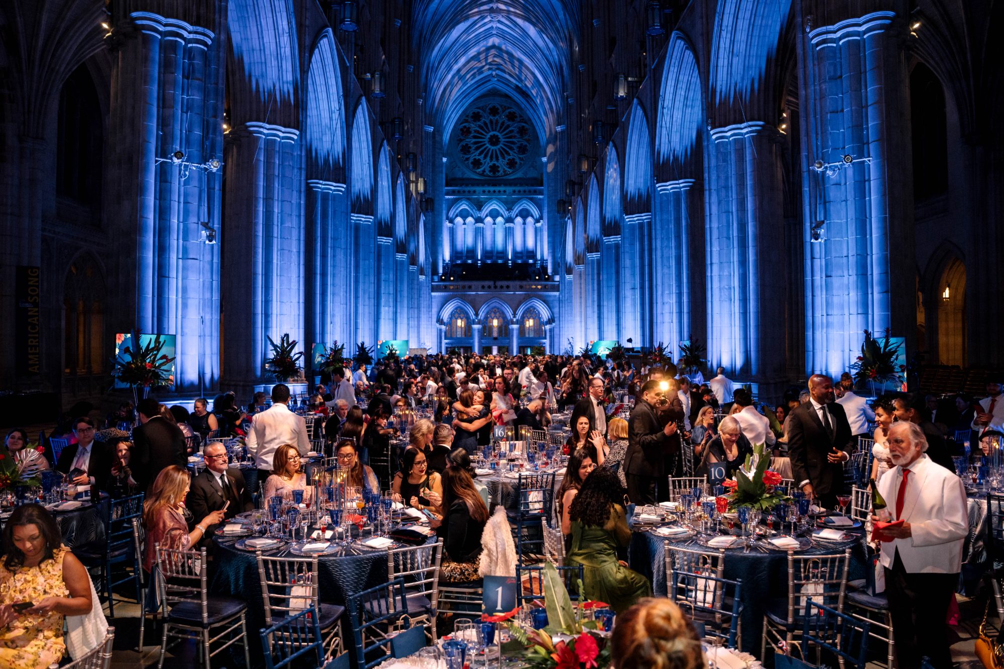 A black-tie gala dinner set beneath the vaulted nave of a cathedral, tables filling the aisle under blue architectural lighting