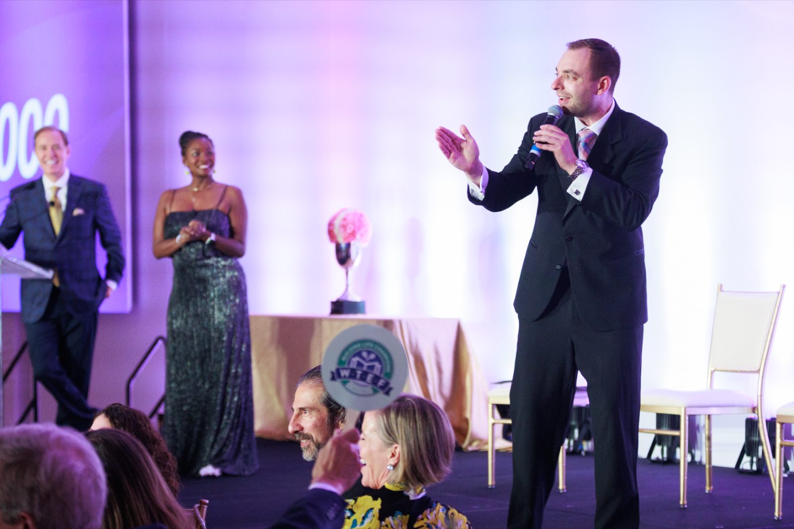 Max Wolf hosting a benefit auction, with honorees on stage and guests laughing in the foreground